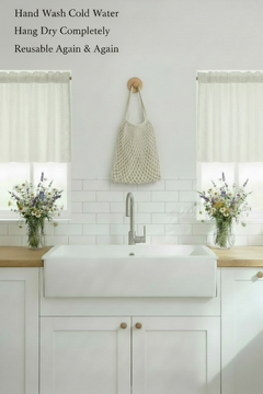 A white farmhouse-style sink with a tall chrome faucet installed in a bright white kitchen cabinet with light wood countertops. Two windows are visible on the wall, and a cream reusable bag hangs from a hook above the sink.