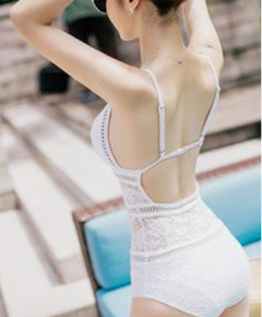 A woman shown from the back wearing a white lace one-piece swimsuit with thin shoulder straps and an open back, standing near a blue lounge chair in an outdoor setting.