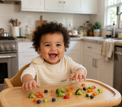 A smiling toddler with curly dark hair sits in a wooden high chair in a bright kitchen, reaching toward colorful baby food pieces on a light wood tray. The child wears a cream and beige striped long-sleeve top and a white bib with a small visible Poppy Baby logo.