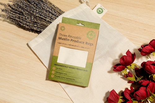 Three reusable muslin produce bags with packaging on a wooden surface with red flowers.